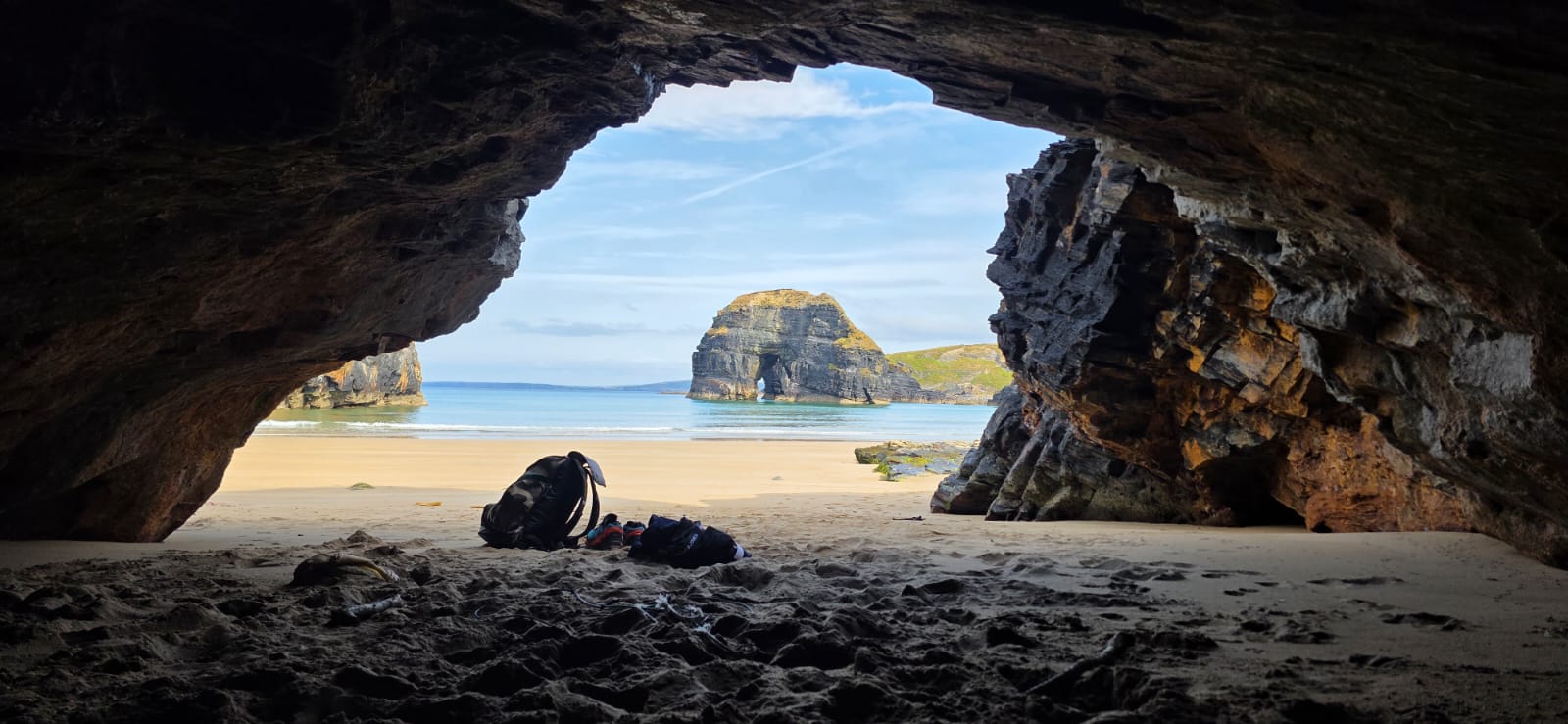 Two rucksacks on the sand inside a sea cave, with a rock arch and turquoise water beyond the opening