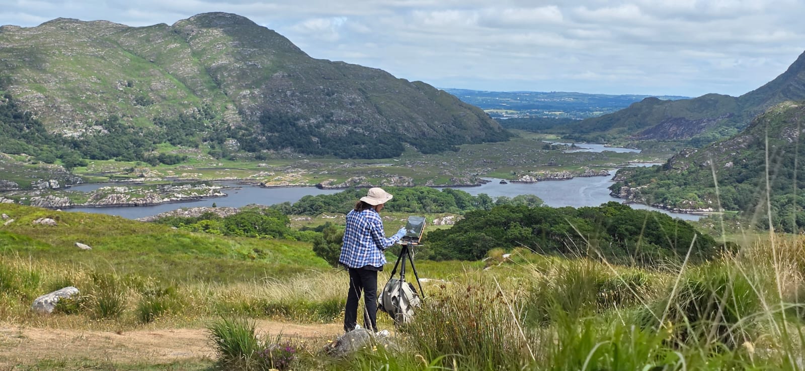 A painter at an easel on a grassy viewpoint above lakes and wooded mountains