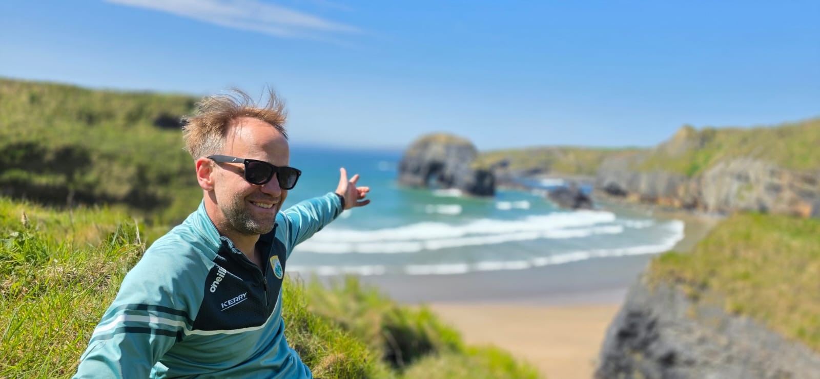 Alexander Schroeder in sunglasses on a grassy clifftop, pointing down at a sandy cove with a sea stack and surf